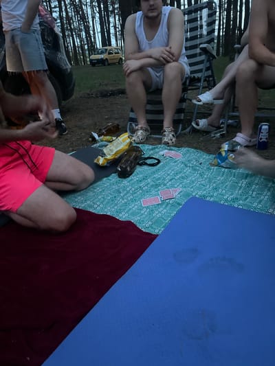 In Uhlyshche, Ukraine, an outdoor gathering takes place in a forested area. Four individuals are present, some seated on chairs and others on blankets spread on the ground. A yellow car is visible in the background, along with a dark-colored vehicle, possibly an SUV. The scene includes a picnic blanket with playing cards, an opened bag of chips, and several bottles. A blue mat with footprints is also present. The lighting suggests it is either dusk or dawn.