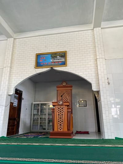 The image captures the interior of a mosque in Blang Bebangka, Indonesia. A wooden pulpit, ornately carved with Islamic motifs, stands prominently in the center, beneath an arched alcove. To the left of the pulpit, a glass-fronted cabinet filled with books is visible. A wooden door is also on the left, and a framed digital clock displaying prayer times hangs on the wall above the alcove. Green carpeting covers the floor, with a smaller prayer rug placed in front of the cabinet. The walls are tiled with white rectangular tiles. The environment appears clean and orderly. There are no people visible in the image.