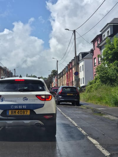 The image shows a street scene in Sanem, Luxembourg. In the foreground, the rear of a white and blue Kia car, registration BM 4087, belonging to paramedicus.lu, a home care and physiotherapy service, is visible. The car is parked on the side of the road. Behind the Kia, there is a black Volvo, registration LT 4007, and another car. A "no parking" sign is placed along the curb. The background features a row of colorful buildings and light poles. The sky is cloudy. The road appears wet.