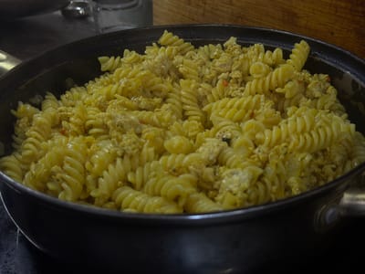 The image shows a close-up, high-angle view of fusilli pasta mixed with scrambled eggs and possibly some small pieces of meat, all served in a dark, round frying pan. The dish is golden yellow in color and appears to be hot. The pan is set on a dark surface, possibly a stovetop. In the background, out of focus, are some glass containers and a wooden surface. The scene is well-lit, with the light source casting subtle shadows on the pasta. The overall impression is that of a home-cooked meal. The image does not provide any information about the city of Crokhovo, Russia, or its location within that city.