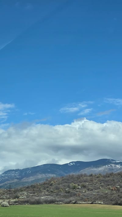 The image shows a landscape under a bright blue sky with scattered cirrus clouds. A large, thick band of cumulus clouds partially obscures the sky towards the middle of the frame. A mountain range is visible, its slopes covered in sparse vegetation and rocky outcroppings, with some snow visible at higher elevations. A green field is present in the lower right corner. The image is oriented vertically. It is not possible to verify the location in the city of Usoika, Bulgaria with the provided information.