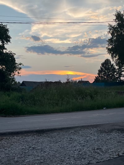The image depicts a rural landscape at dusk, likely in Ludorvay, Russia. The sky is filled with a mix of clouds, with streaks of orange and yellow from the setting sun visible on the horizon. Power lines stretch across the upper portion of the frame. In the middle ground, there are several structures, including a dark-roofed building and a light-colored fence. Dense green vegetation covers the lower middle ground, with a pile of dark material to the left of the building. A paved road runs across the foreground, bordered by a section of loose gravel. Trees frame the left and right sides of the image.