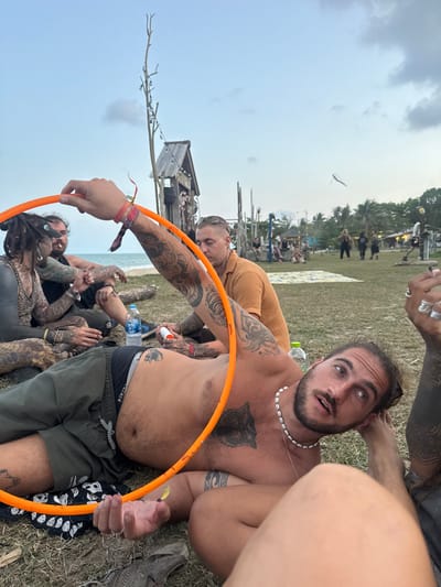 The scene depicts an outdoor gathering in Ban Pang Pring, Thailand, set on a grassy area adjacent to a beach under a light blue, cloudy sky. In the immediate foreground, a shirtless man with a beard, dark hair pulled back, and a shell necklace lies on his right side on the grass, looking upward towards the viewer. He wears dark green shorts over black underwear and displays extensive tattoos, including a large wolf design on his chest/abdomen and tribal patterns on his arms. He holds an orange hula hoop with his left hand, which also contains a small yellow object. An arm belonging to another individual, heavily tattooed and adorned with multiple rings and wristbands, also grasps the hula hoop on the left.
In the midground, several individuals are visible lounging or seated on the grass. One man with short, styled hair and an orange-brown short-sleeved shirt is seated, leaning back, also with tattooed arms. To his left, another man with dreadlocks and extensive dark tattoos is seated, holding a lit cigarette. A plastic water bottle and a white tube are placed on the grass near these individuals. The sandy beach and ocean are visible behind them.
The background features various rustic, artistic wooden structures scattered across the landscape. These include a tall, bare wooden pole strung with lights and a larger, hut-like wooden structure with a peaked roof. Further back, more people are present, standing and sitting. Palm trees line the distant horizon, and a white kite or similar object is visible high in the sky to the right. The overall environment suggests a casual, festive outdoor event by the sea.