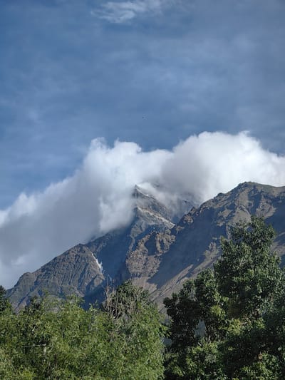 A telephoto shot depicts the rugged, barren slopes of a mountain range partially obscured by dramatic, billowing white clouds. The mountain peaks are sharp and angular, with visible patches of snow clinging to crevices and higher elevations. The lower slopes are textured with a rough, rocky surface, hinting at erosion and geological formations. In the foreground, dense, lush green trees with full foliage create a stark contrast to the starkness of the mountains, their leaves appearing vibrant in the sunlight. The sky above is a clear blue, punctuated by wispy clouds that suggest fair weather, though the prominent clouds around the mountain peaks indicate potential atmospheric conditions at higher altitudes. The scene is natural and untamed, suggesting a remote or elevated landscape. The image does not contain any people or visible man-made structures, focusing entirely on the natural elements of the mountain environment.