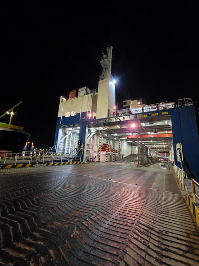 An outdoor, low-angle, wide shot captures the interior deck of a large ferry or RoRo (Roll-on/Roll-off) ship at night, docked in the port of Ravenna, Italy. The foreground displays a textured, dark-colored deck with a chevron pattern, showing signs of wear. Yellow and black striped safety barriers line the right side of the deck, leading towards the rear of the vessel.

The midground reveals the open interior of the ship, illuminated by overhead lights. A ramp extends upwards and into the ship's interior. Several levels of the ship's structure are visible, with various pipes, lights, and mechanical components. Red and white striped barriers are present, along with orange traffic cones and some signage, including one that reads "ZUCCO CARGO." The overall impression is one of industrial infrastructure, designed for the transport of vehicles and cargo. In the background to the left, the dark hull of another ship is partially visible. The sky above is completely black.