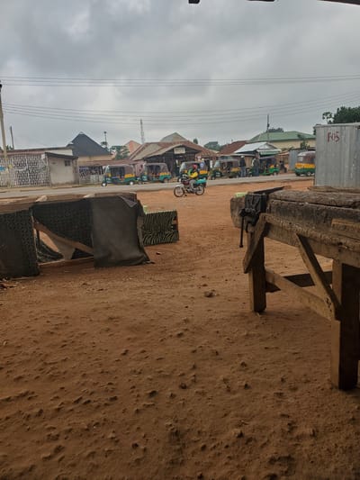 The media file depicts a scene in Barikin Sabon Gidan Kanat, Nigeria, on an overcast day. The primary subjects are a row of colorful auto-rickshaws, locally known as "keke napep," parked along a dirt road. One keke, in yellow and green, has "PTA 1057" visible on its side. Another, in purple and green, shows "PTA 1011." In the foreground, a rustic wooden workbench with a vise attached is positioned on the dusty ground. Adjacent to it are some makeshift seating arrangements, including a low structure covered in dark fabric and a smaller item with a zebra-like pattern.
The scene appears to be a transportation hub or a waiting area. The presence of the parked kekes suggests that people are either waiting for passengers or have just completed a journey. The overcast sky indicates it might be morning or late afternoon, with no direct sunlight. The setting is characterized by dusty, reddish-brown earth and a mix of simple buildings and structures, including what looks like a shipping container with "POS" written on it in red. Power lines crisscross the sky, a common feature in many urban and semi-urban areas. No specific human activities are captured in motion, but the overall impression is one of a typical day in a Nigerian community where kekes are a vital mode of transport.