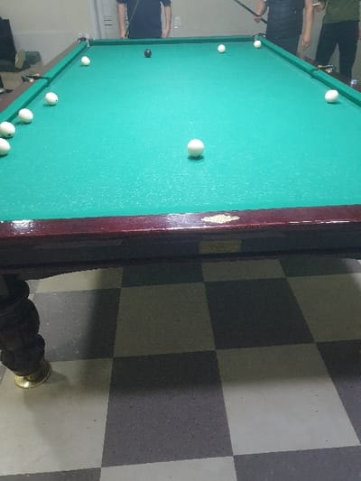 A top-down shot reveals a billiards table with a green felt surface and several white balls scattered across it, along with one black ball. The table has a dark wooden frame with ornate detailing. Part of the floor surrounding the table is visible, showcasing a checkered pattern of alternating dark and light squares. Some people are visible on the far left.