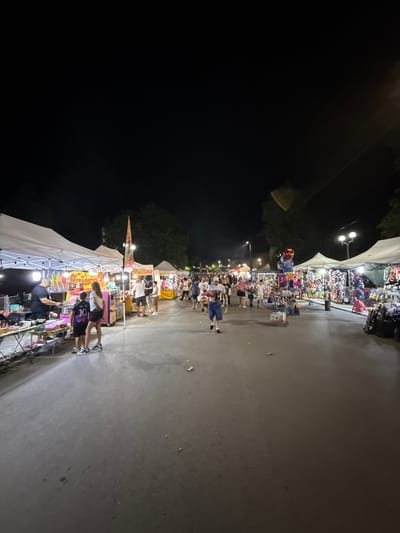 The image depicts a night scene at an outdoor market or fair, likely in the city of Stob, Bulgaria. The scene is illuminated by artificial lights from the stalls and streetlights, creating a lively atmosphere. The market extends along a paved walkway, with numerous tents and stalls set up on either side. These stalls appear to be selling a variety of goods, including food items like ice cream and cotton candy, as well as toys and souvenirs.
Several people are visible, walking along the path, browsing the stalls, and interacting with vendors. Some are dressed casually, indicating a relaxed evening outing. The overall impression is one of a community gathering or a popular tourist attraction. The background shows trees and the dark night sky, suggesting an urban or semi-urban setting. The ground surface is asphalt or a similar paved material.