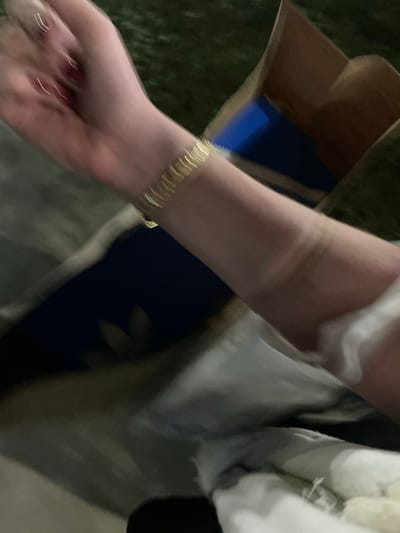 A person's arm is shown with a gold bracelet on the wrist. There is a visible tan line on the arm. The person is holding something out of frame and there is a beige bag in the background. The person appears to be wearing a light colored shirt or jacket. The picture is blurry.