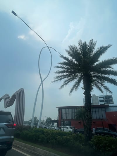 The image depicts an outdoor scene in the city of Medan, Indonesia, likely during daylight hours under an overcast sky. In the foreground, a grey SUV is parked or moving slowly on a road, with its rear visible. To the left, a large, abstract sculpture with vertical striped elements is present. A tall, white, looping sculpture also stands prominently in the frame. A large palm tree with dark green fronds is situated in the mid-ground, obscuring part of a modern building with a red facade and large windows. The building appears to be a commercial establishment. Several other vehicles are visible in the background, along with a multi-story building under construction. A few individuals are sparsely scattered near the building. The overall environment suggests a busy urban area with some architectural points of interest and vegetation.
