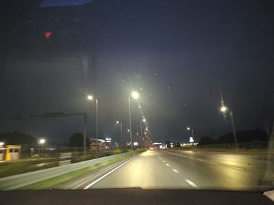 The image depicts a nighttime view from inside a vehicle traveling on a highway, likely on the outskirts of Laagri alevik, Estonia. The sky is dark and overcast, with streetlights illuminating the road ahead. The highway appears to be multi-lane, with white lines marking the lanes. On either side of the road, there are grassy embankments and trees, with some buildings and structures visible in the distance. The overall impression is one of a typical highway scene at night, with the road and its surroundings lit by artificial lights.