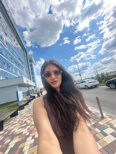A woman with long dark hair and sunglasses is taking a selfie on a city street. In the background, there is a tall, modern building with glass windows and a blue sky with fluffy white clouds. There are parked cars on the side of the street, along with benches and potted plants. The ground is paved with colorful bricks. Based on these details, the location appears to be the city of Gavrilkovo, Russia, near a modern office building or residential complex.
