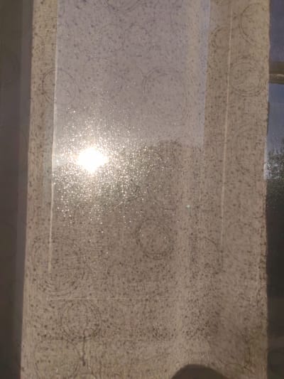 The image depicts a close-up view of a translucent, patterned curtain, likely made of light fabric, hanging in front of a window. The sun is shining brightly through the curtain, creating a dazzling effect with numerous small, sparkling highlights. This suggests it is daytime, and the sun is either rising or setting, given the warm, golden light.

The curtain has a subtle, repeating pattern of concentric circles and small dots, which are visible through the diffused sunlight. The fabric itself appears to be a light, neutral color, possibly beige or off-white. The sunlight is intense enough to cause some glare, making it difficult to discern details outside the window clearly. However, hints of trees and possibly a building can be seen through the fabric on the right side of the image, indicating an outdoor scene beyond the window.

The overall mood is serene and warm, evoking a sense of peacefulness associated with natural light. The focus is on the interplay of light and fabric, with the sparkling effect of the sunlight catching the texture of the material. No people or specific objects are prominently featured, the subject being the natural phenomenon of light filtering through a domestic setting. There is no visible text in the image. The location context of Сокол, Russia, provides a geographical setting for this scene of everyday life.