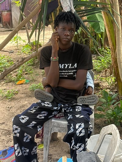 A young Black person with dark, curly hair is seated on a gray plastic chair in an outdoor environment. The individual is wearing a black t-shirt with white lettering that reads "MYLA" and has a bracelet on their left wrist. Their legs are crossed, and they are wearing patterned black and white pants and gray slip-on shoes. A baby, wearing a light blue long-sleeved shirt, is being held in the person's lap, with one of the baby's feet visible. The background consists of dirt, scattered green plants, and the trunks of trees. Behind the person, a purple building is visible, suggesting an urban or semi-urban setting. The scene appears to be in Tawpo, Nigeria, as indicated by the specific location.