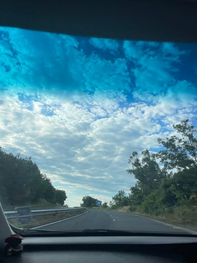 The image captures a view from inside a vehicle traveling on a road that appears to be a highway or a major route. The road is paved and marked with white lines. On either side of the road are lush green trees and vegetation, suggesting a rural or semi-rural environment. The sky is a prominent feature, filled with dramatic white and gray clouds against a bright blue sky, indicating it might be a partly cloudy day with good visibility. The foreground shows the dashboard of the vehicle, with a small decorative item that resembles a cowboy figurine with a hat. In the distance, there are signs and a few vehicles, suggesting the presence of other traffic. There is a guardrail visible along the side of the road. The overall impression is of a journey through a scenic landscape under a dynamic sky. The user prompt mentions the city of Balabanovo, Bulgaria, but there are no specific visual cues within the image to confirm this location.