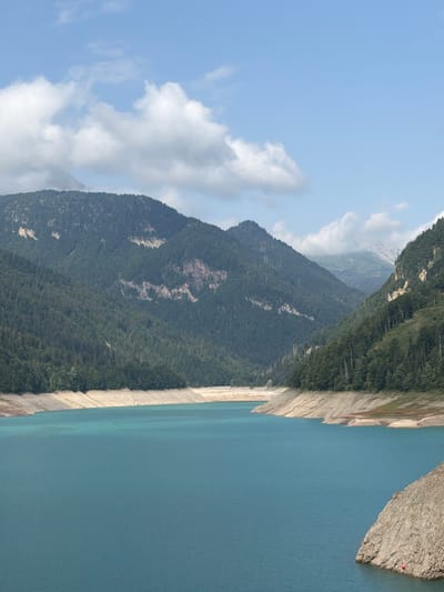 This is a vertical, high-angle shot of a vast, turquoise lake in a mountainous region. The lake occupies the lower two-thirds of the frame, with its surface calm and reflective. The shores of the lake are characterized by steep, rocky slopes with exposed, dry earth and scattered vegetation. In the background, towering mountains covered in dense green forests rise towards a partly cloudy blue sky. The overall impression is one of natural grandeur and a remote, pristine landscape. The scene is uninhabited by people. The location is the Ampezzo/Dimpeç area of Italy.