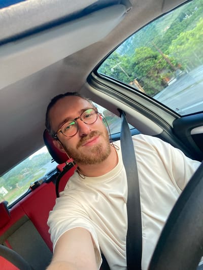 A man with fair skin, a reddish-brown beard, and round glasses is in the driver's seat of a car. He is wearing a white shirt and has a seatbelt across his chest. The car's interior is gray and red. Through the car window, green trees and glimpses of buildings can be seen. The image appears to be taken from a low angle, looking up and slightly angled. The location is Đurmani, Montenegro.