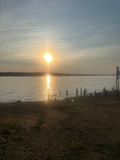 This image depicts a sandy beach scene at the city of Ust-Kachka, Russia, during sunset. The sun is low in the sky, casting a bright, shimmering reflection on the calm water of a lake or river. A silhouetted treeline forms the distant horizon. Several people are present on the beach and in the shallow water, some appearing to be swimming or wading. Beach chairs and other objects are scattered on the sand. The foreground is dominated by the textured sand of the beach.