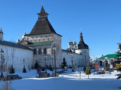 This image depicts a snow-covered winter scene within a fortified historical complex in Kozlovo, Russia, under a clear blue sky. A tall, light-colored stone tower with a dark, pointed wooden roof and a small cupola dominates the left side of the frame, featuring arched windows and a green architectural band. Adjacent to it, a high, white crenellated stone wall extends leftward, behind which silver onion domes with crosses are visible. Further along the wall to the right, another white tower with a dark, onion-shaped dome and a pointed finial is observed.
The foreground and midground consist of a snow-covered courtyard or park area. Multiple dark-colored park benches are partially covered by the snow, and several slender, bare deciduous trees along with small evergreen conifers are planted throughout. Numerous black, multi-lantern streetlights are strategically placed. A single individual, dressed in dark clothing, is visible walking on the snow in the midground, moving towards the right side of the frame. To the far right, a building with a green roof and a white, arched facade is partially visible. Bright sunlight casts distinct shadows across the snow.