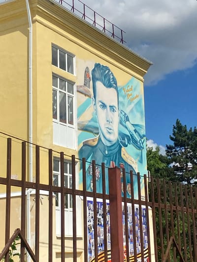 The image shows a mural painted on the side of a yellow building in Akhtyrskiy, Russia. The mural depicts a portrait of a man in military uniform, identified by Russian text on the mural as "Чубисов Юрий Васильевич". He is shown with a fighter jet in the background. Below the portrait, a series of smaller portraits are displayed, along with what appear to be ribbons or medals. The building has several windows and is partially obscured by a dark metal fence in the foreground. Trees are visible behind the building. The sky is blue with some clouds.