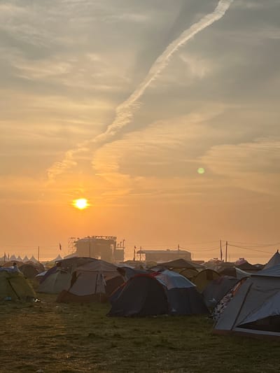 The image depicts a campground at sunrise in Surakovo, Russia. The sky is a warm orange and yellow hue, with scattered clouds and a prominent contrail stretching across the upper portion of the frame. The sun is visible as a bright orb just above the horizon, casting a golden light over the scene.
In the foreground, numerous tents of various colors and sizes are pitched on a grassy field. The tents appear to be made of fabric and are set up for camping. Some tents are partially open, revealing glimpses of their interiors. In the background, a large stage structure is visible, suggesting the campground may be associated with an outdoor event or festival. Beyond the stage, a hazy landscape stretches to the horizon. The overall atmosphere is serene and quiet, characteristic of early morning in a camping area.