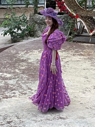 A person stands in front of a textured, light-colored wall with distressed patterns. The individual is wearing a striking purple dress with a polka dot pattern of pink dots. The dress has voluminous, puffy sleeves and a wide brimmed hat adorned with matching purple ruffles. A red belt cinches the waist of the dress. The person is smiling and looking to their left, with their left hand resting on their dress, wearing a white bracelet.
Behind the person, to the left, is a lush green plant with visible branches and leaves. In the background, there is a large, intricate structure made of woven branches, decorated with red flowers and illuminated by a string of warm white lights. Parts of what appear to be stone walls and possibly windows are visible behind the foliage and the woven structure.
The image is oriented vertically, and the lighting suggests it could be daytime, possibly outdoors, given the natural elements present. The overall scene evokes a whimsical or celebratory atmosphere. There is no visible text in the image. The location context is Nong Prue, Thailand.