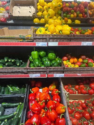 The image captures a vibrant fresh produce section, likely within a grocery store in Noisy-le-Grand, France. The scene features abundant displays of colorful vegetables neatly arranged in woven baskets, cardboard boxes, and black plastic crates.
The primary subjects are various fresh vegetables. Bright yellow bell peppers are prominent on the left, some filling woven baskets and others overflowing from a cardboard box. Above them, a diverse collection of mixed tomatoes in shades of red, orange, yellow, and green are visible. Green bell peppers are showcased in baskets in the middle section, while below them, dark green zucchini are stacked in plastic crates. On the right, numerous red and orange bell peppers are displayed alongside clusters of red tomatoes on the vine.
No people are visible, focusing the attention entirely on the produce. The setting is clearly a retail environment, with clear pricing and origin information for the goods.
Notable details include the organized presentation by color and type, the fresh appearance of the vegetables, and the distinct containers. An empty cardboard box with an avocado graphic is also visible. The lighting is bright and even, suggesting an indoor setting.
Visible text from the price labels includes:
- "POIVRON JAUNE PAYS BAS Le kg 4,99€" (Yellow bell pepper from the Netherlands, 4.99€ per kilogram).
- "TOMATE MELANGEES FRANCE Le kg 6.50€" (Mixed tomatoes from France, 6.50€ per kilogram).
- "POIVRON ROUGE PAYS BAS Le kg 4,99€" (Red bell pepper from the Netherlands, 4.99€ per kilogram).