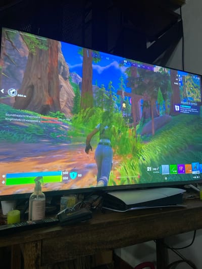 The image depicts a close-up indoor scene, likely in a residence in San Antonio de Padua, Argentina, focused on a television displaying a video game. A large, dark-framed flat-screen television dominates the upper half of the frame, showing gameplay from what appears to be Fortnite. The game screen displays a female character with brown hair, wearing a dark vest and light blue jeans, running on a dirt path through a lush, forested environment with large trees, green foliage, and distant rock formations under a bright blue sky. User interface elements visible include health and shield bars (100/100 and 50 shield respectively), a kill feed in Spanish indicating "Soundthealarm18 eliminó a crazykachori con un rifle" and "KingKoala82 (2) escopeteó a JewelOfWisdom (1)", a mini-map, a compass pointing NE (93 degrees), and weapon slots at the bottom right showing various items and ammunition counts (e.g., shotgun with 240 ammo, assault rifle with 187 ammo). An "ANOMALÍA ACTIVA" (Active Anomaly) notification in Spanish is also visible on the right.

Below the television, on a rustic dark wooden table, are several items. To the right, a white and black PlayStation 5 console is positioned horizontally. To the left, a clear plastic bottle of hand sanitizer with a white pump is visible, next to a small green cylindrical candle. A black TV remote control and a dark wallet or pouch with white earbuds or cables resting on it are situated between the hand sanitizer and the console. The area directly above the television shows a dark, textured ceiling or wall structure.
