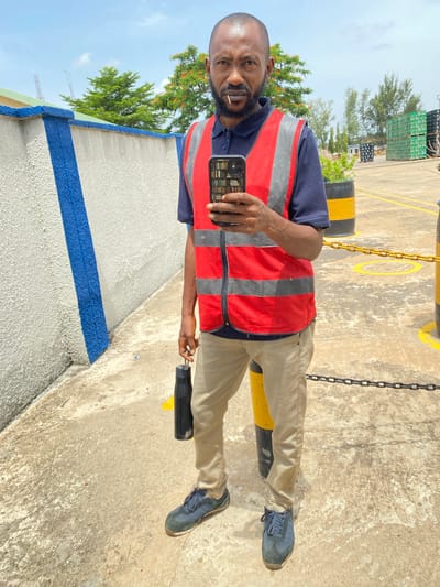 The image shows a man in an outdoor industrial setting in Kinkinau, Nigeria. The man is wearing a dark blue collared shirt, a red and gray high-visibility vest, and tan pants. He has a beard and is looking at the camera, holding a smartphone in his right hand. He appears to be taking a selfie. He is also holding a small object, possibly a pen, in his left hand. A black water bottle with a yellow stripe is attached to his pants.
The background includes a concrete ground, some yellow and black striped barriers, and chains. Further in the distance, there are stacks of what appear to be crates, trees, and construction cranes, suggesting a working environment such as a port, warehouse, or construction site. The sky is visible and appears partly cloudy, indicating it is daytime. The lighting suggests a sunny day. The man's expression is neutral. There is no visible text in the image.