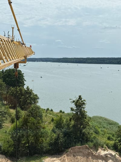 This is a landscape photograph taken outdoors on a bright, cloudy day. The image is oriented vertically. In the foreground, a yellow construction crane extends from the lower left corner upwards and towards the right. The crane appears to be assembled and ready for use, with its lattice structure clearly visible. Above the crane, the sky is a pale blue with scattered white clouds.
The middle ground of the image features a wide body of water, possibly a lake or a bay. Several small white sailboats are dotted across the surface of the water, suggesting a recreational or sporting activity. The water is calm and reflects the light from the sky. Along the far edge of the water is a dense, dark green line of trees, forming a natural barrier.
The right side of the image shows a steep, rocky embankment covered with lush green vegetation and trees. The textures of the rocks are rough and varied, with some areas appearing lighter and others darker. The trees are dense and leafy, creating a vibrant green contrast with the pale water and sky.
The overall impression is one of a serene natural setting with a touch of human industry. The bright sky and calm water suggest pleasant weather, likely during daylight hours. The presence of sailboats hints at activities like sailing or boating. The rocky embankment and dense foliage indicate a natural landscape, possibly a lakeside or coastal area. No text is visible in the image.