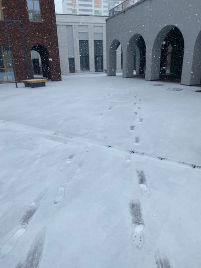 The image shows a snow-covered ground or path in an urban environment. There are footprints in the snow, indicating recent passage. Buildings are visible on one side, featuring architectural elements like arches and windows. The buildings' exterior materials include brick and concrete. It's snowing. Location information: Chemoshur, Russia.
