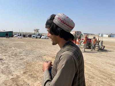 The image depicts an open, unpaved area in Pir Dust, Afghanistan, under a clear blue sky. In the immediate foreground, a young man is positioned on the right side of the frame, viewed from his right, facing left. He has dark hair and wears a white cap with red patterned embroidery, a light brown long-sleeved shirt, and a dark brown plaid vest. He holds a blue object in his right hand. His mouth is slightly open. The ground is dry, composed of light brown soil and small stones.
In the midground to the left, a group of approximately ten individuals, some dressed in traditional white Afghan attire, are congregated near several parked white vehicles, including SUVs. A green and blue tent is partially visible on the far left, and a light-colored building with a green roof stands in the distant background. To the right, two utility tractors are present. Three individuals stand near them; two men are wearing orange high-visibility vests over their clothing, and a woman in a headscarf is also visible. Additional structures are discernible on the distant horizon.