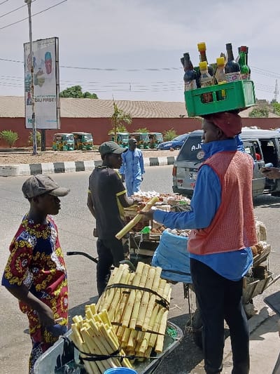 The image depicts a street scene in Communauté rurale de Payar, Senegal, under an overcast sky. In the foreground, three individuals are engaged in commerce on the side of a paved road.
To the left, a young male wearing a multicolored patterned short-sleeved shirt and a dark baseball cap stands next to a black pushcart containing several bundles of peeled sugarcane stalks. He faces right, observing the central figures.
In the center, a person wearing a blue long-sleeved shirt, an orange mesh vest, and a red cap is positioned with their back partially to the viewer, holding a green plastic crate filled with various bottled beverages balanced on their head. This individual stands behind a second pushcart that also contains sugarcane and other goods. Their right arm extends forward, holding out a single peeled sugarcane stalk.
To the immediate left of this central figure, partially obscured, a second male wearing a dark t-shirt with yellow accents on the sleeves and a dark baseball cap is present, facing the central figure. He appears to be either receiving or handing off the sugarcane stalk.
In the midground, across the street, several green and yellow auto-rickshaws (tuk-tuks) are parked in a row near a reddish-brown building with a low wall. A male in a light blue traditional garment walks past these vehicles, moving right to left.
Further back, a large billboard with a political advertisement featuring two men and text (partially visible, "EAU FIRST!") stands next to the road. The background consists of distant trees, structures, and power lines.