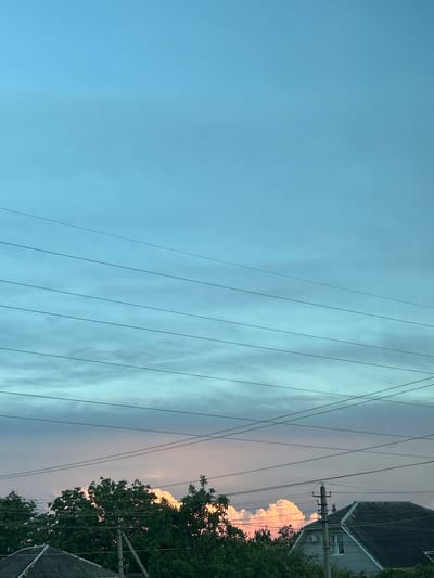 The image displays an outdoor scene, likely a residential or suburban area, looking upwards towards the sky. The left side of the sky is a clear, light blue, gradually transitioning to a cloudier, grey-blue hue towards the right. Numerous thin, dark power lines stretch horizontally across the frame, cutting through the sky. Some lines appear parallel, while others intersect, connecting to utility structures.

On the far right, parts of two residential building roofs are visible, along with a wooden utility pole featuring multiple crossarms and insulators. Below the structures and pole, a vibrant green canopy of tree foliage occupies the lower right corner. Behind the trees and houses, a cluster of clouds glows with warm pink and golden light, strongly indicating either sunrise or sunset. The light conditions suggest the "golden hour" of the day. No people or discernible text are present. The scene is a static sky view, typical of a populated area, set in Imeni Maksima Gor'kogo, Russia.