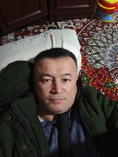 A man lies on a patterned rug with a white pillow propped behind his head. He is wearing a green jacket over a blue patterned shirt. The rug features a mix of geometric and floral designs in red, brown, white, and black. A small, colorful plastic bucket sits in the upper right corner near dark wooden cabinets. The man's face is the focal point, looking directly at the camera with a neutral expression.