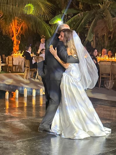 The image shows a bride and groom dancing in what appears to be a wedding reception. The bride is wearing a white wedding gown and veil, while the groom is in a dark suit. The scene appears to be outdoors, with lush tropical vegetation (possibly palm trees) visible in the background. The lighting suggests it is either late afternoon or evening, with some candles and possibly string lights illuminating the area. There are wedding guests seated at tables in the background. The setting appears to be in the Communauté rurale de Malicounda, Senegal based on the environment.