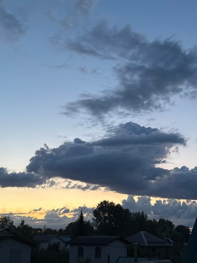 This image depicts a twilight sky over a residential area in Volzhsk, Russia. The sky is a gradient of pale blue at the top, transitioning to a warm yellow and orange horizon where the sun has recently set. Dramatic, dark gray cumulus clouds dominate the upper and middle sections of the sky, with lighter, wispy clouds scattered below near the horizon.
The lower portion of the image shows the rooftops of several houses, characterized by dark gray and dark blue shingles. Some houses have visible chimneys. The structures appear to be of modest size, typical of a suburban or rural setting. Trees, primarily dark green and silhouetted against the sky, form a dense line across the mid-ground, obscuring the lower half of most buildings. A few taller coniferous trees stand out among the deciduous ones. The overall impression is one of a quiet evening in a Russian town as daylight fades. There are no people or animals visible in the image.