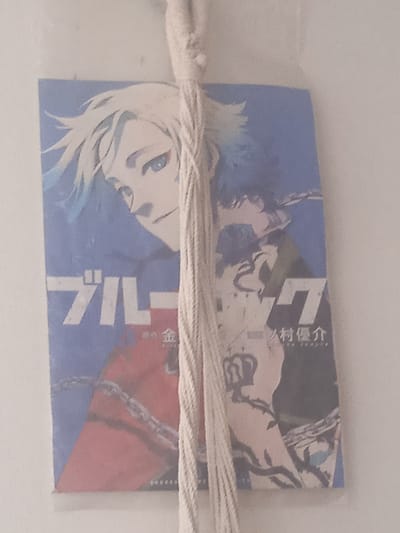 A vertical, slightly blurry photograph captures a hanging, rectangular item, likely a bookmark or a small poster, displaying anime-style artwork. The artwork features a character with white hair that transitions to blue at the tips and striking blue eyes. The character's face is turned to the left, with a faint smile. The background is predominantly a gradient of blue. The bottom portion of the artwork is divided diagonally, with the left side appearing red and featuring a stylized chain, and the right side featuring the same character in a more detailed pose, wearing a dark outfit and a crown-like symbol. Japanese text is visible on the artwork, with "ブルーロック" prominently displayed. The item is suspended by a thick, cream-colored cord. The background of the photograph is a textured, off-white surface, indicative of an interior wall. The image was taken in Puerto La Cruz, Venezuela.
