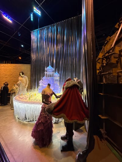This media file captures a nighttime scene, likely indoors, possibly a studio or a display area. The primary subjects are mannequins dressed in elaborate costumes, suggesting a theatrical or retail display. One mannequin is adorned in a dramatic red cape with fur trim, reminiscent of royalty or a festive character. Another is draped in a voluminous, layered purple gown. Further down, a mannequin wears a patterned, lighter-colored garment. The background features a visually striking installation made of translucent, possibly ice-like structures illuminated with cool blue and purple lights, creating a magical or wintery atmosphere. Behind this, a metallic, ridged curtain adds to the theatrical backdrop. In the upper portion of the image, industrial-looking structures and scaffolding are visible, indicating a backstage or work-in-progress environment. The lower part of the scene shows a floor with visible scuff marks, suggesting it's a functional space. Outside the immediate display, strings of colorful lights, some in red, blue, and yellow, are visible against a dark sky, hinting at an external light display or decorative lighting. There is no visible text in the image. The overall impression is one of a festive or dramatic presentation, with a focus on costumes and a fantastical, illuminated setting.