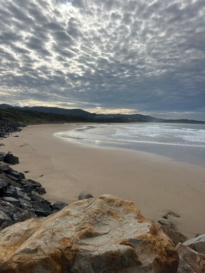 The image depicts a cloudy day at a beach, likely in Coffs Harbour, Australia. The foreground is dominated by large, light-colored rocks, some with reddish-brown staining, leading down to a sandy beach. Darker, jagged rocks form a breakwater along the left side of the frame. The beach stretches to the right, meeting a gentle ocean with small waves. In the mid-ground, a few people are scattered along the shoreline. The background features a tree-covered coastline with some buildings visible, culminating in rolling hills under a dramatic sky filled with textured, grey clouds, with patches of bright light suggesting the sun is attempting to break through.
