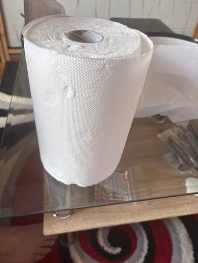 The image shows a roll of white paper towels resting on a glass-topped table. A partial view of a rug with a circular red, grey, and black pattern is visible to the right of the table. A person's foot is visible near the corner of the rug. The location is unknown, and therefore cannot be confirmed to be Mirkovo, Bulgaria.