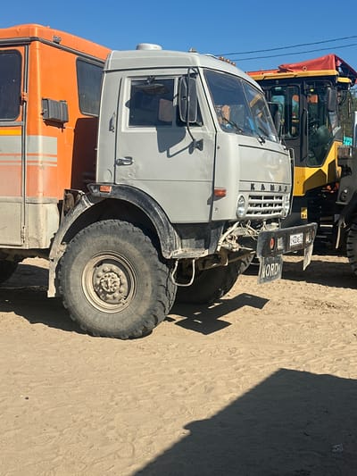 The image shows two vehicles parked on a sandy surface under a clear blue sky. The foreground is dominated by the front portion of a gray truck with an orange truck body behind it. The gray truck has large, rugged tires, a prominent front grille with the word "KAMAZ" visible, and a black license plate with the characters "C432AE 11". Below the license plate, another black plate reads "NORD". A portion of a yellow construction vehicle with a red canopy is visible to the right of the gray truck. The environment appears to be an outdoor area, possibly a construction site or a storage yard, with sand covering the ground and tire tracks visible. No people are clearly depicted in the scene, though a reflection in the gray truck's window suggests the presence of at least one person inside. The lighting suggests it is daytime, with strong sunlight casting shadows. The location is identified as Chikshino, Russia.