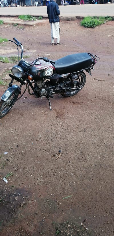 A black and red motorcycle is parked on a dirt road in Kassa Market, Nigeria. The motorcycle has a black seat and a luggage rack on the back. A circular decal on the side of the motorcycle reads "MC 79". In the background, several people are standing on a paved road. One person in the background is wearing a blue jacket and a long white skirt. The scene appears to be outdoors during the day, with overcast weather suggested by the lighting. The ground is a mixture of dirt and gravel, with some patches of wet mud. There is some sparse green grass growing along the edges of the road. The overall atmosphere is that of a busy marketplace.