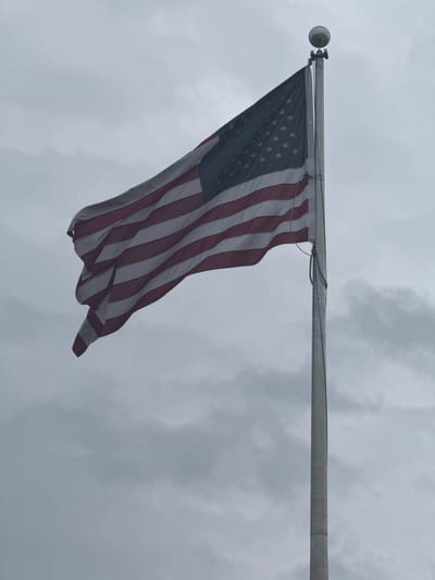 The image shows an American flag hanging from a flagpole against an overcast sky. The flag is waving in the wind, displaying the stars and stripes. The flagpole is a gray metal pole with a spherical top. The environment appears to be outdoors, possibly in Mountain Park, United States of America, based on the prompt. No people or interactions are visible in the image.