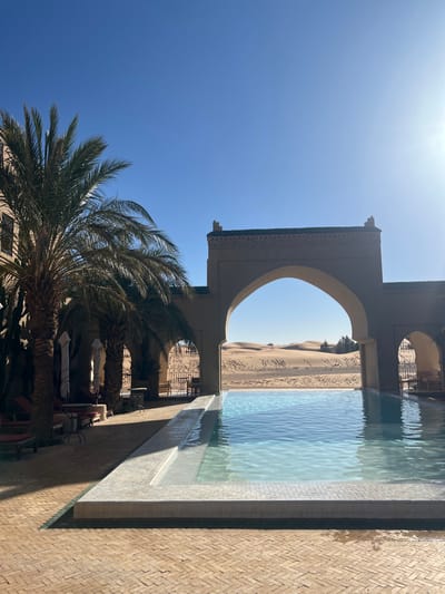 The image displays an outdoor scene at a resort or hotel in Rissani, Morocco, bordering a desert. In the foreground, a brick-patterned patio leads to a light blue swimming pool with white-tiled edges, appearing to have an infinity design. To the left of the pool, several palm trees cast shadows over an area furnished with red lounge chairs, small tables, and white umbrellas, adjacent to a light-colored building with dark windows. In the midground, a prominent tan-colored archway, topped with dark green decorative tiles, frames a clear view of vast sand dunes in the background. A smaller archway is partially visible to the right. The sky above is clear and bright blue, with the sun's glare visible in the upper right. No individuals are present in the scene.