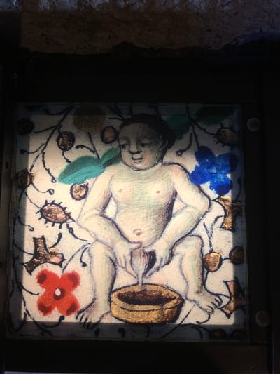 An image of a tile depicting a medieval illustration is visible. The tile is set in a dark frame, possibly a display case or mounted on a wall. The illustration depicts a nude figure with fair skin in a reclined pose, centered on a white or cream-colored background. The figure is surrounded by floral designs, including a blue flower, a red flower, and stylized leaves, all interconnected by black vines or stems. The tile is located in the Château de Chillon, a castle on the shores of Lake Geneva, in the commune of Veytaux, Switzerland.