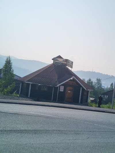 The image shows a log cabin-style building with a sign that reads "СУВЕНИРЫ" (Souvenirs) on its roof. The building has a steeply pitched, dark-colored roof and log walls with white decorative elements around the windows and corners. There is an orange-colored door on the front of the building, and a smaller sign with text in Russian is visible above it.
To the left of the main building, there is a tall green tree. In the background, rolling hills covered in light blue mist can be seen, suggesting a mountainous or hilly landscape. Power lines are visible crisscrossing the sky. A dark-clothed person is standing to the right of the building, facing away from the camera. The foreground is dominated by a gray asphalt road with a white line marking. The overall scene appears to be a roadside establishment in a somewhat rural or mountainous area. The presence of the "Souvenirs" sign indicates it is likely a place of commerce catering to tourists. Based on the architecture and the likely context of a souvenir shop in a scenic area, this image is likely located in the vicinity of the ski resort town of Sheregesh, Russia, known for its natural beauty and mountainous terrain.