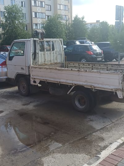 In Elitnyi, Russia, a white flatbed truck is parked on a paved surface near a multi-story apartment building. The truck appears weathered, with visible rust on its bed and other components. A spare tire is secured underneath the truck bed. Other vehicles, including dark-colored cars and SUVs, are parked behind the flatbed truck. Trees line the area, and the sky is overcast. The ground surface shows signs of wetness, possibly from recent rain, creating reflections of the surrounding environment.