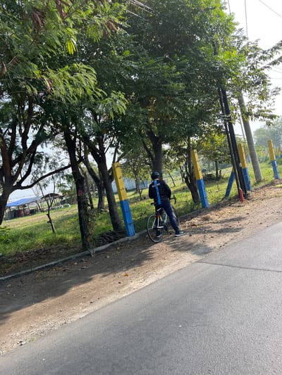 A person is standing next to a bicycle on the side of a paved road in Gempolsari, Indonesia. The person, wearing a black helmet and a black long-sleeved shirt with blue and white stripes on the back, is facing away from the camera and looking towards a grassy area with trees. The bicycle is a road bike with a white frame and orange accents. In the background, there are several poles painted yellow and blue, a fence, and a blue tarpaulin structure. The environment appears to be a rural or semi-rural area with abundant greenery and clear daylight. The road surface is asphalt.