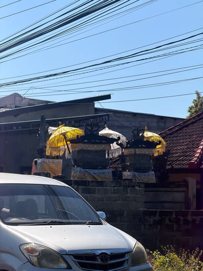 The image shows a street scene in Kubu, Indonesia, with a silver minivan in the foreground. Behind the minivan, there are several traditional Balinese temple umbrellas, some yellow and some white, set up on stone structures. The umbrellas are decorated with intricate patterns and tassels. In the background, there are buildings with tiled roofs and a brick wall. Overhead, a tangle of power lines stretches across the bright blue sky. The scene suggests a place of cultural significance, possibly during a festival or ceremony.